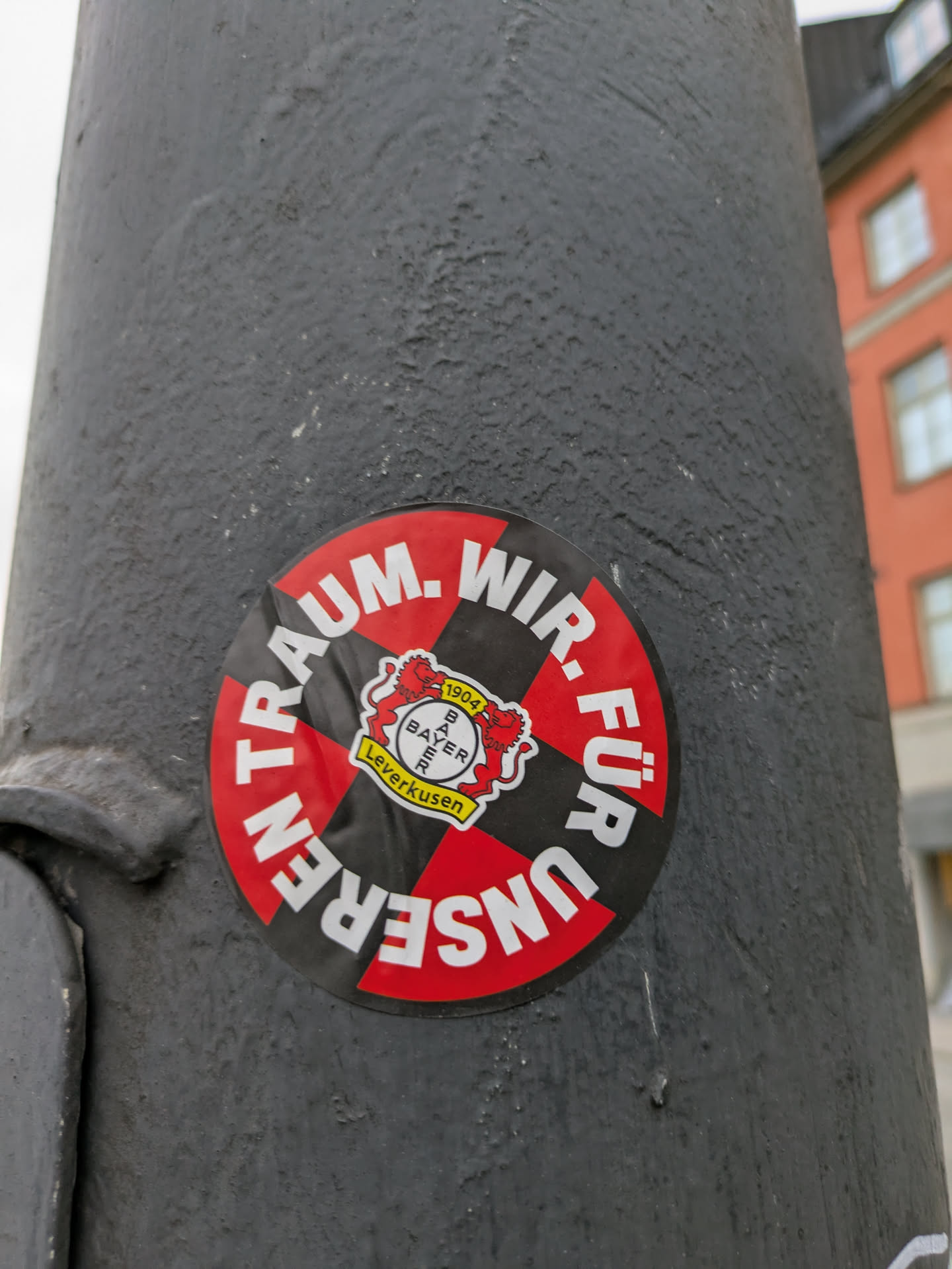 A round sticker featuring the Bayer Leverkusen logo and the text "Wir für unseren Traum" (We for our dream) in German. The sticker is black and red with a central logo depicting two lions and the year 1904. The sticker is affixed to a gray pole outdoors.