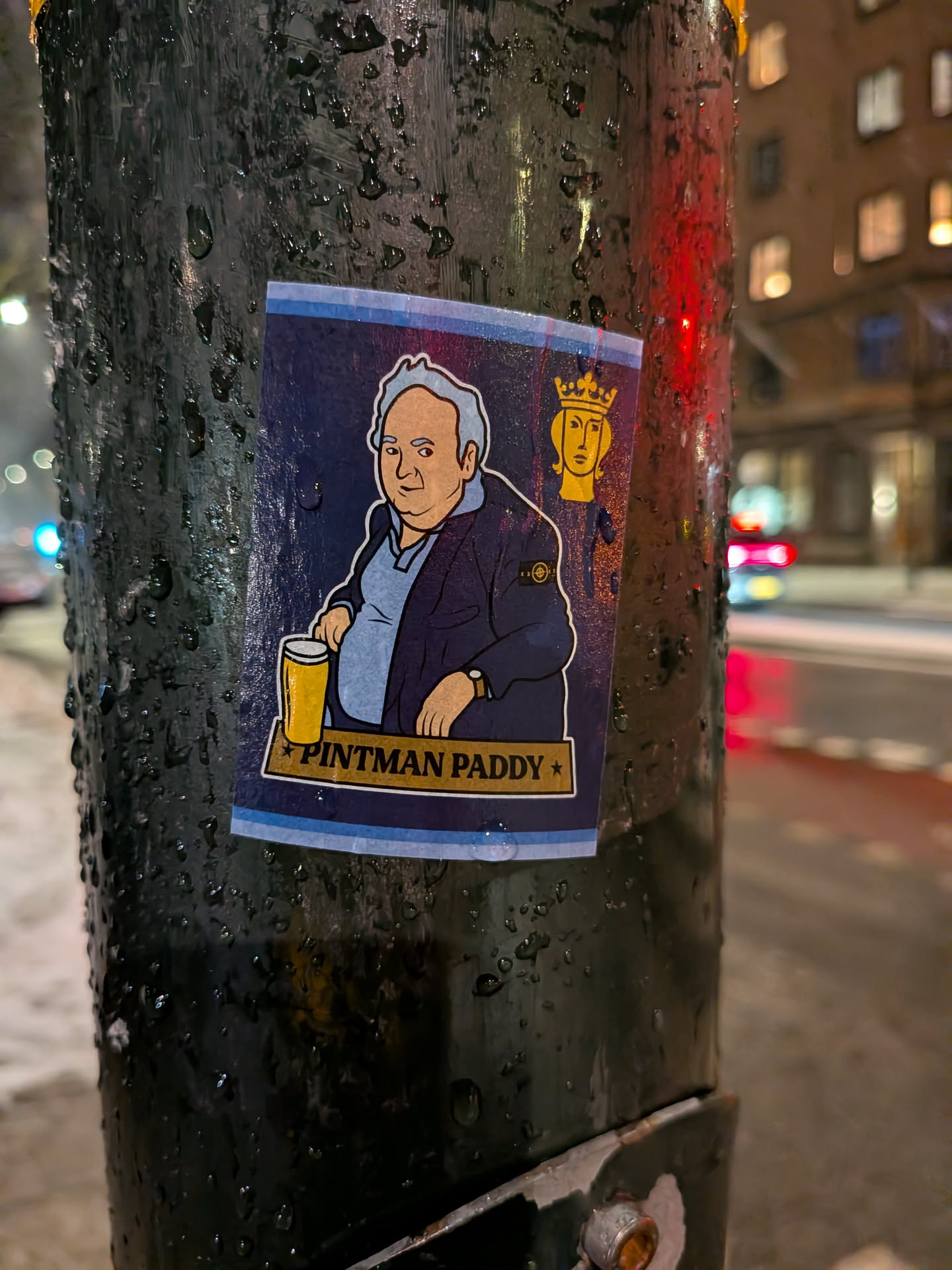 The sticker is a vertically oriented rectangle with a dark blue background. It features a cartoon illustration of a middle-aged, slightly portly man with light grey hair and fair skin, wearing a light blue polo shirt and a dark blue jacket. On the left sleeve of his jacket, there is a circular badge resembling the Stone Island compass logo in yellow and black. The man holds a tall glass of amber-colored beer (a pint) in his right hand. To the right of his head, a golden, stylized head wearing a crown is depicted. Below the man, a golden banner displays the text 'PINTMAN PADDY' in dark letters, flanked by two small golden stars. The bottom edge of the sticker has a light blue and white striped border. The sticker is applied to a dark, wet pole, with visible water droplets.