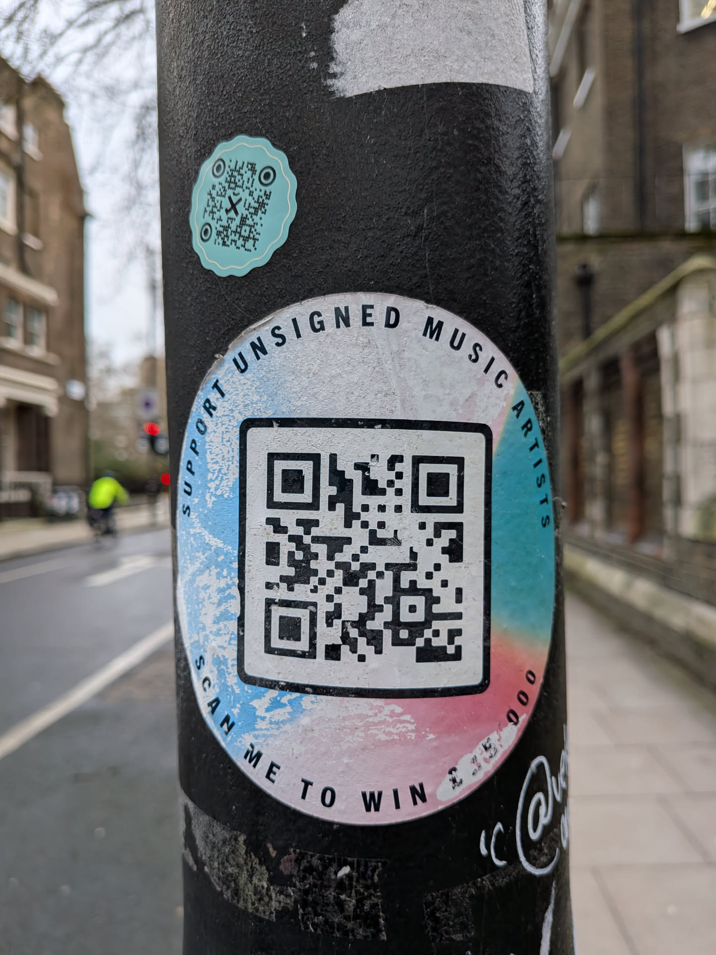 A circular sticker adhered to a black pole, featuring a large black QR code prominently in its center against a white background. The border of the sticker displays a gradient of light blue on the left and top, transitioning to a soft pink on the right and bottom. Curved black text encircles the QR code. The sticker shows signs of wear, with some scuffing and fading on the colored border. Above this main sticker, there is a smaller, teal-colored, scalloped-edge sticker, also containing a QR code with a small 'X' symbol in its center.