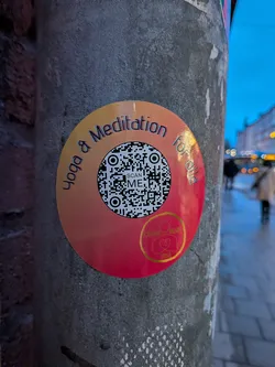 The sticker is circular, featuring a vibrant gradient background that transitions from a warm orange at the top to a rich red at the bottom. Centered on the sticker is a black and white QR code, beneath which the words "SCAN ME" are printed. Arcing around the top edge of the circle, in a playful, slightly shadowed font, are the words "Yoga & Meditation for all". In the bottom right quadrant, there's a smaller circular logo with a golden outline, containing two intertwined heart-like shapes and the text "GOLDEN HEART YOGA" in gold. The sticker is affixed to a grey, textured pole.