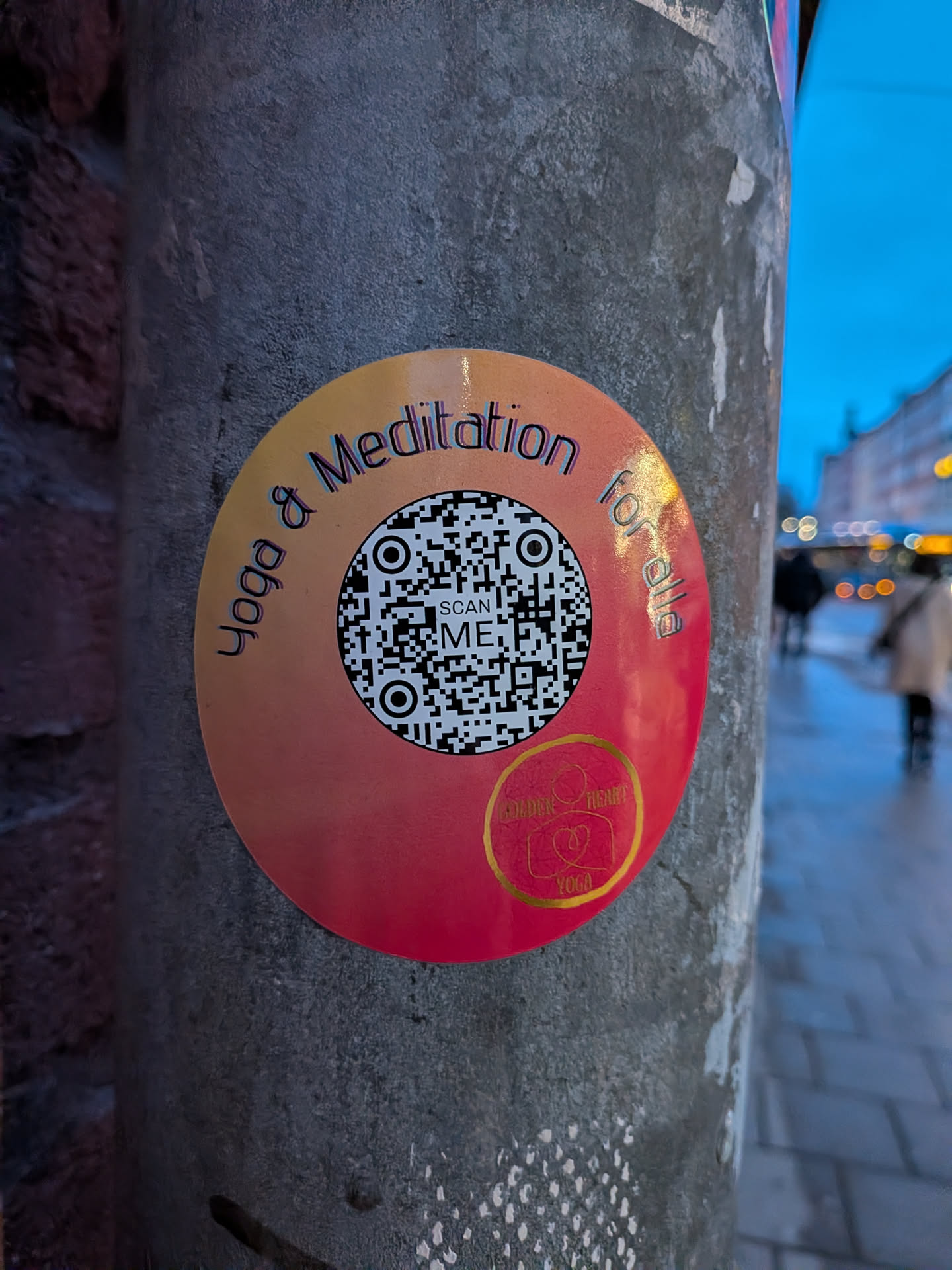 The sticker is circular, featuring a vibrant gradient background that transitions from a warm orange at the top to a rich red at the bottom. Centered on the sticker is a black and white QR code, beneath which the words "SCAN ME" are printed. Arcing around the top edge of the circle, in a playful, slightly shadowed font, are the words "Yoga & Meditation for all". In the bottom right quadrant, there's a smaller circular logo with a golden outline, containing two intertwined heart-like shapes and the text "GOLDEN HEART YOGA" in gold. The sticker is affixed to a grey, textured pole.