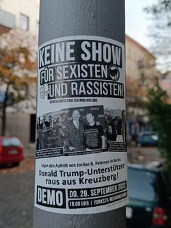 A black and white sticker on a pole protesting against Jordan B. Peterson's  show in Berlin, calling him a supporter of Donald Trump. It promotes a demo on September 29th, 2022 at 6 PM.