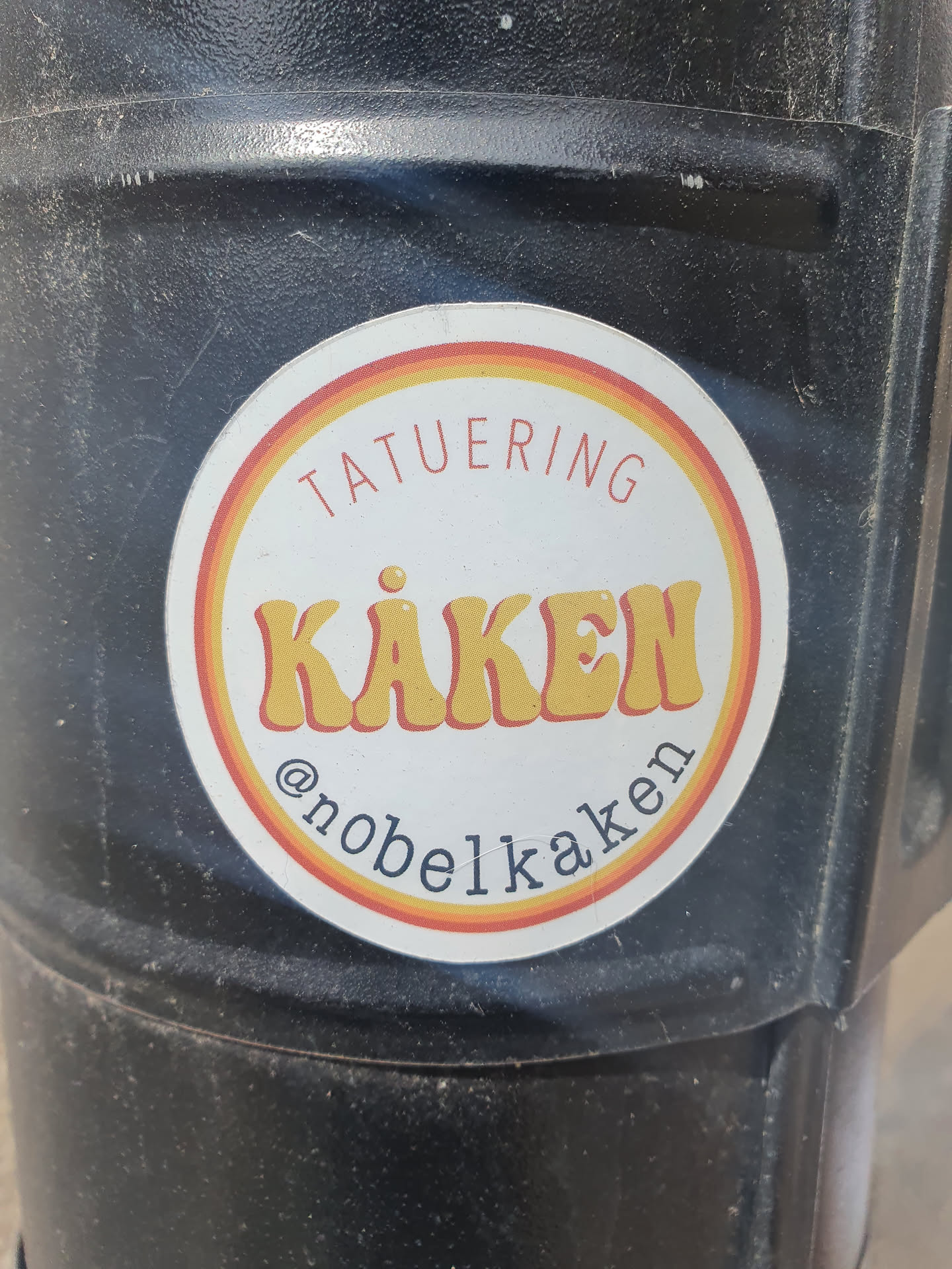 Round sticker with orange, yellow and red stripes.  It says TATUERING (tattoo) in the upper part and KÅKEN in a retro style font in the middle. At the bottom, it shows the Instagram handle @nobelkaken.