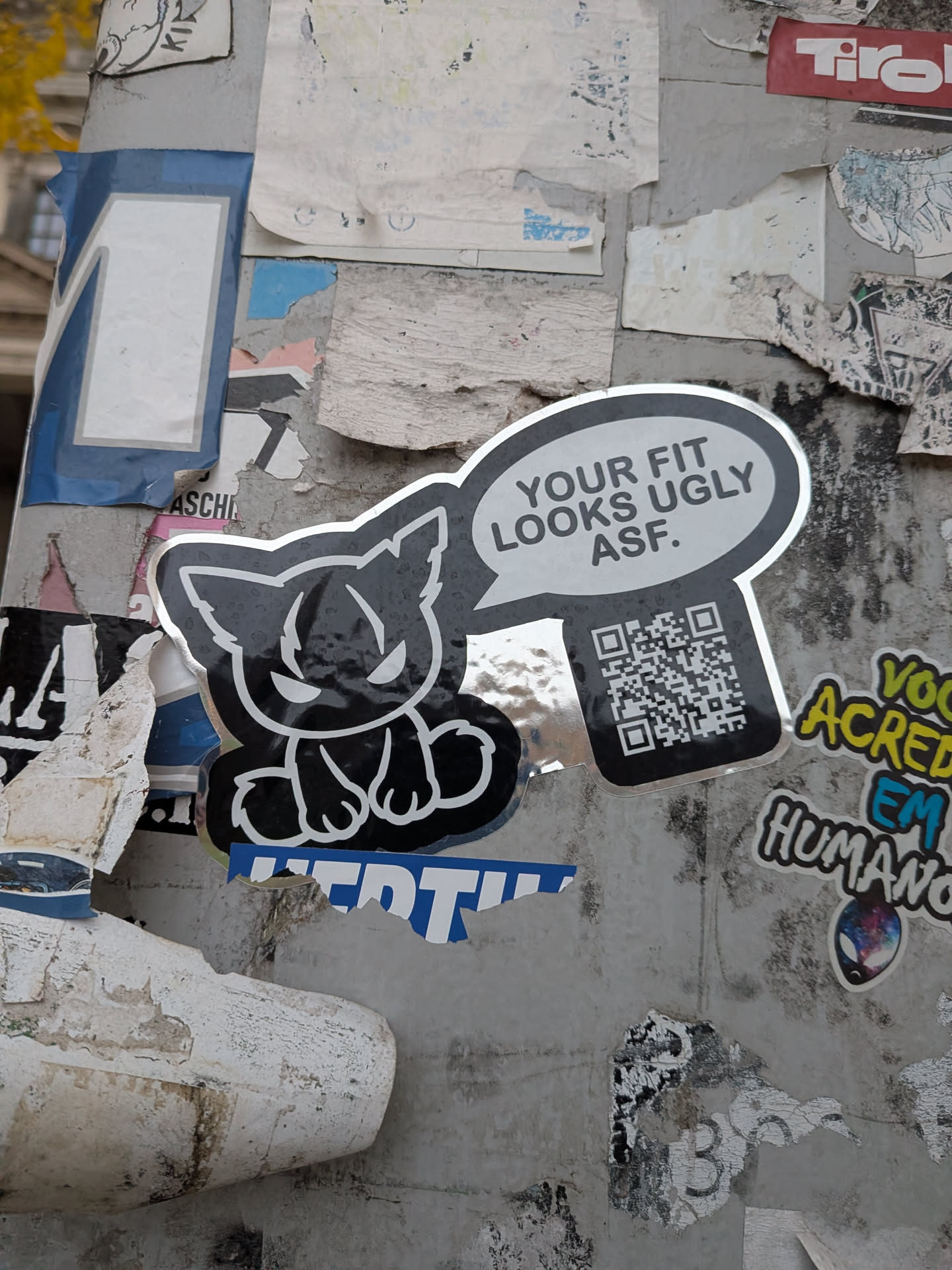 A die-cut sticker with a shiny silver foil border depicting a stylized, grumpy black cat sitting down with sharp, angry eyes. A large speech bubble next to the cat reads 'YOUR FIT LOOKS UGLY ASF.' in bold black uppercase letters, and directly underneath the speech bubble is a prominent QR code set against a dark background.