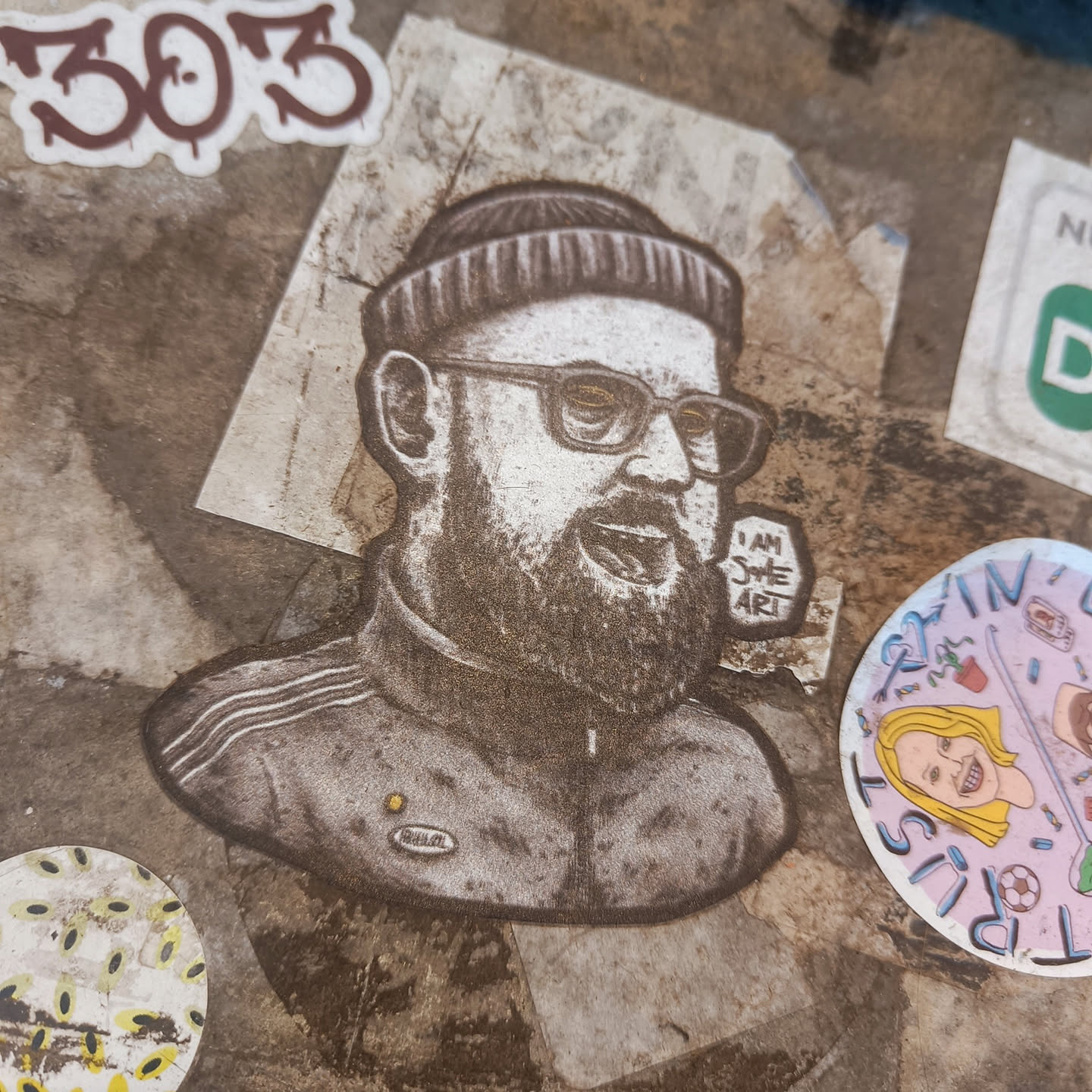 This sticker features a stylized black and white illustration of a man's upper body. He has a beard, glasses, and is wearing a beanie and a zip-up jacket.  The style is reminiscent of printmaking or etching. Small text on the sticker says "I AM SHE ART".