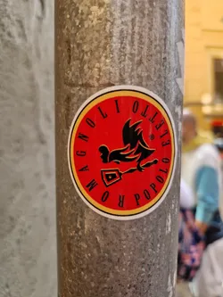 A round sticker with a red background and yellow border.  It features a black illustration of a rooster holding a tool, possibly a shovel. The text "ROMA POPOLO" is written around the rooster in a circular design.