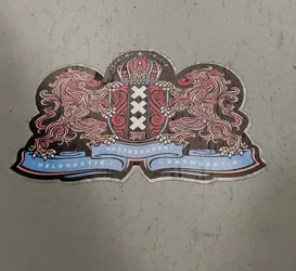 A sticker depicting the coat of arms of Amsterdam.  The design features two lions supporting a central shield with the Amsterdam cross and the year 1940. The ribbon at the bottom reads "Heldhaftig" and "Barmhartig". The top of the shield includes a crown and the letters "O.H.M. O.I.B."