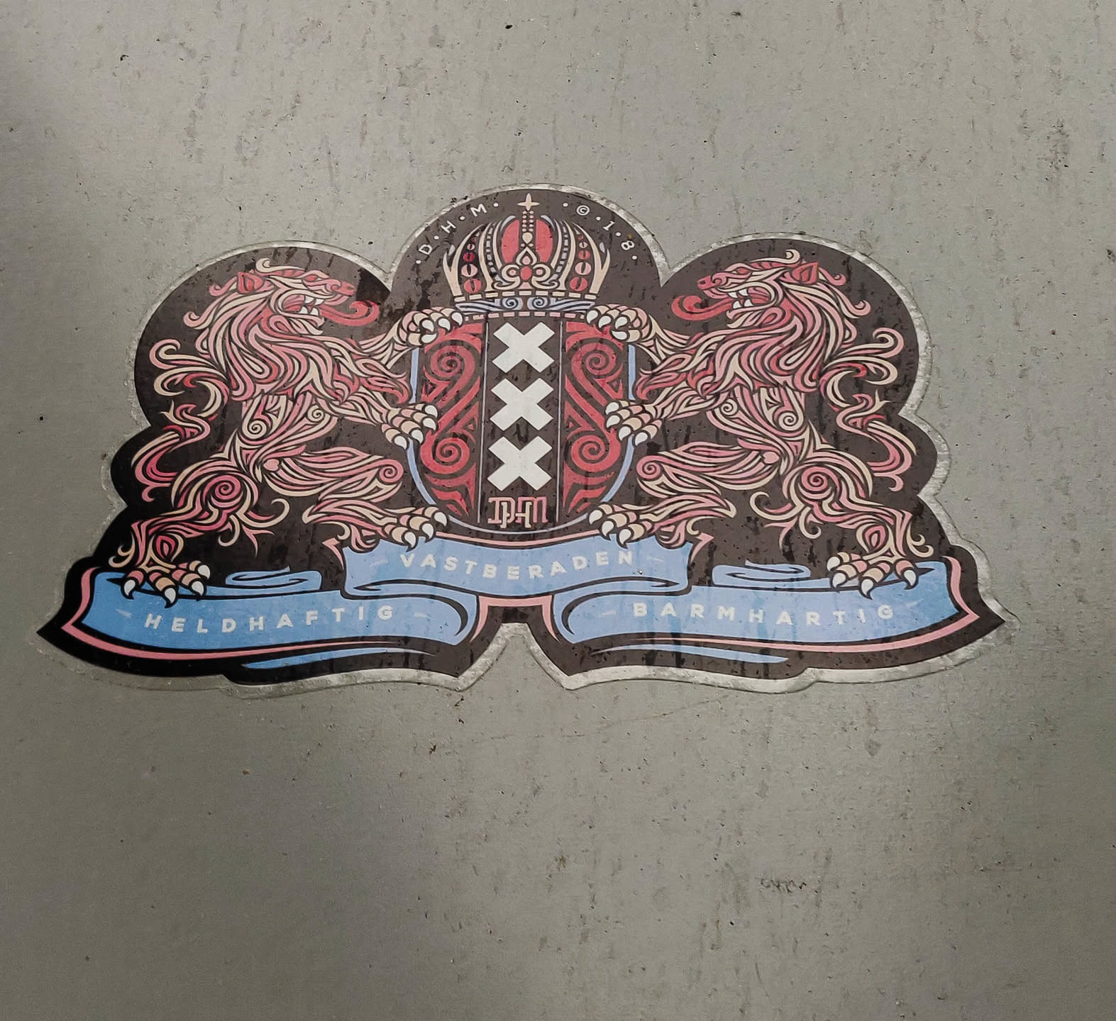 A sticker depicting the coat of arms of Amsterdam.  The design features two lions supporting a central shield with the Amsterdam cross and the year 1940. The ribbon at the bottom reads "Heldhaftig" and "Barmhartig". The top of the shield includes a crown and the letters "O.H.M. O.I.B."