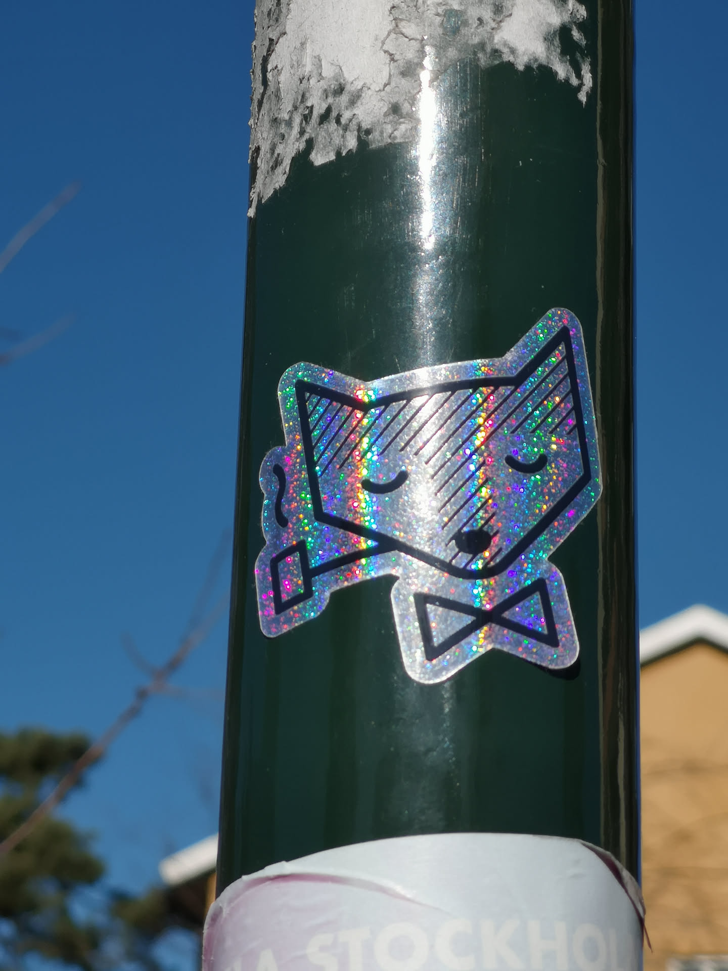 The sticker features a stylized animal head, resembling a fox or a cat, with pointed ears, closed eyes, and a small, serene smile. It is depicted wearing a black outline bow tie below its chin and holding a small pipe in its left paw (viewer's right). The entire surface of the sticker is holographic, reflecting light in a vibrant, glittery spectrum of rainbow colors including pinks, blues, greens, yellows, and purples. All the character's features, the bow tie, and the pipe are outlined with thick black lines, creating a distinct contrast against the shimmering background. The sticker is applied to a dark green pole.
