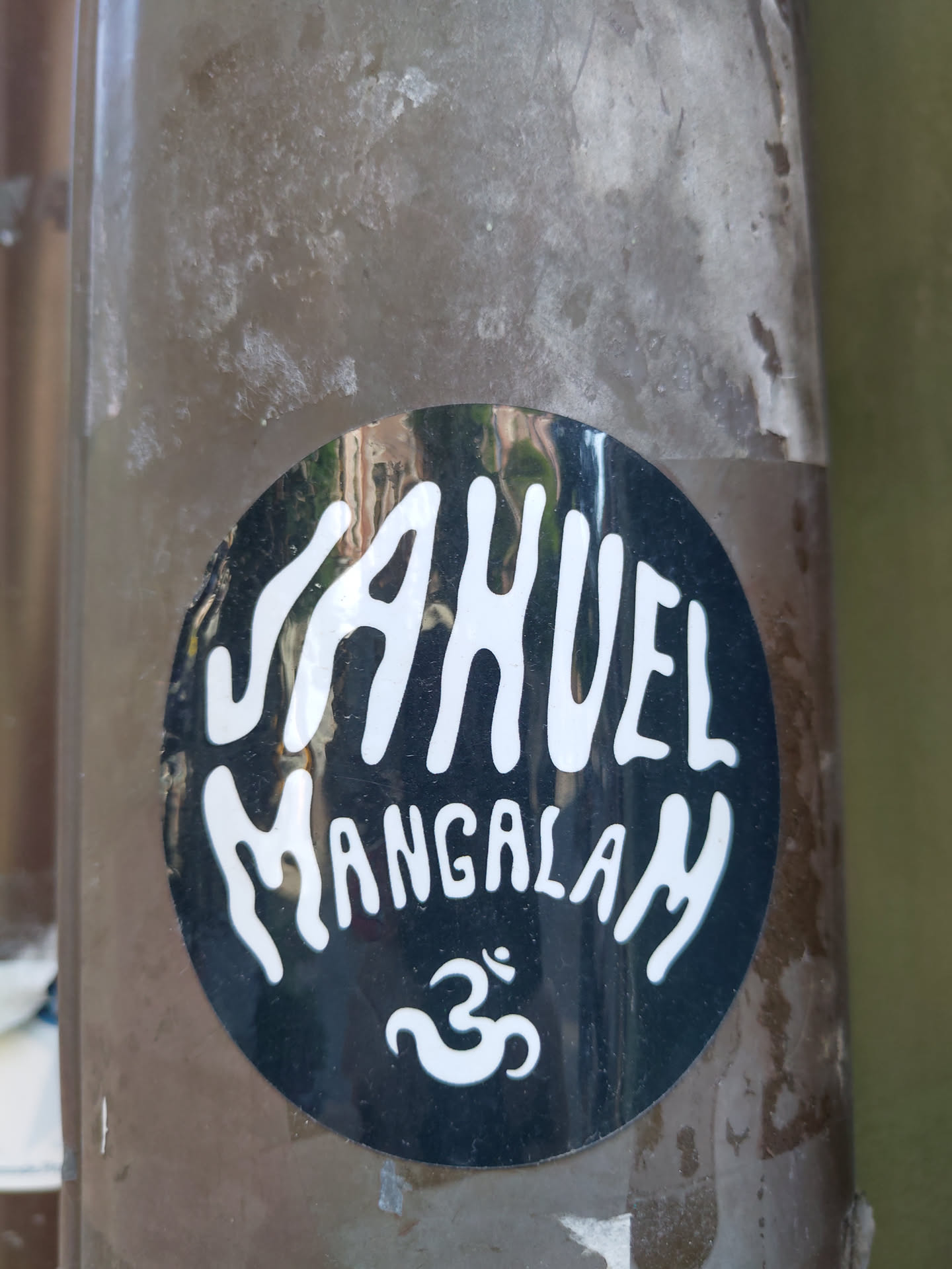 A round black and white sticker with the text JAHUEL MANGALAM and the Om symbol. The sticker is attached to a brown pole.