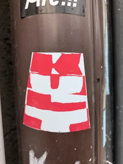 A close-up shot of a red and white abstract sticker on a brown surface. The sticker appears to be hand-made or screen-printed.  There is no discernible text or QR code.