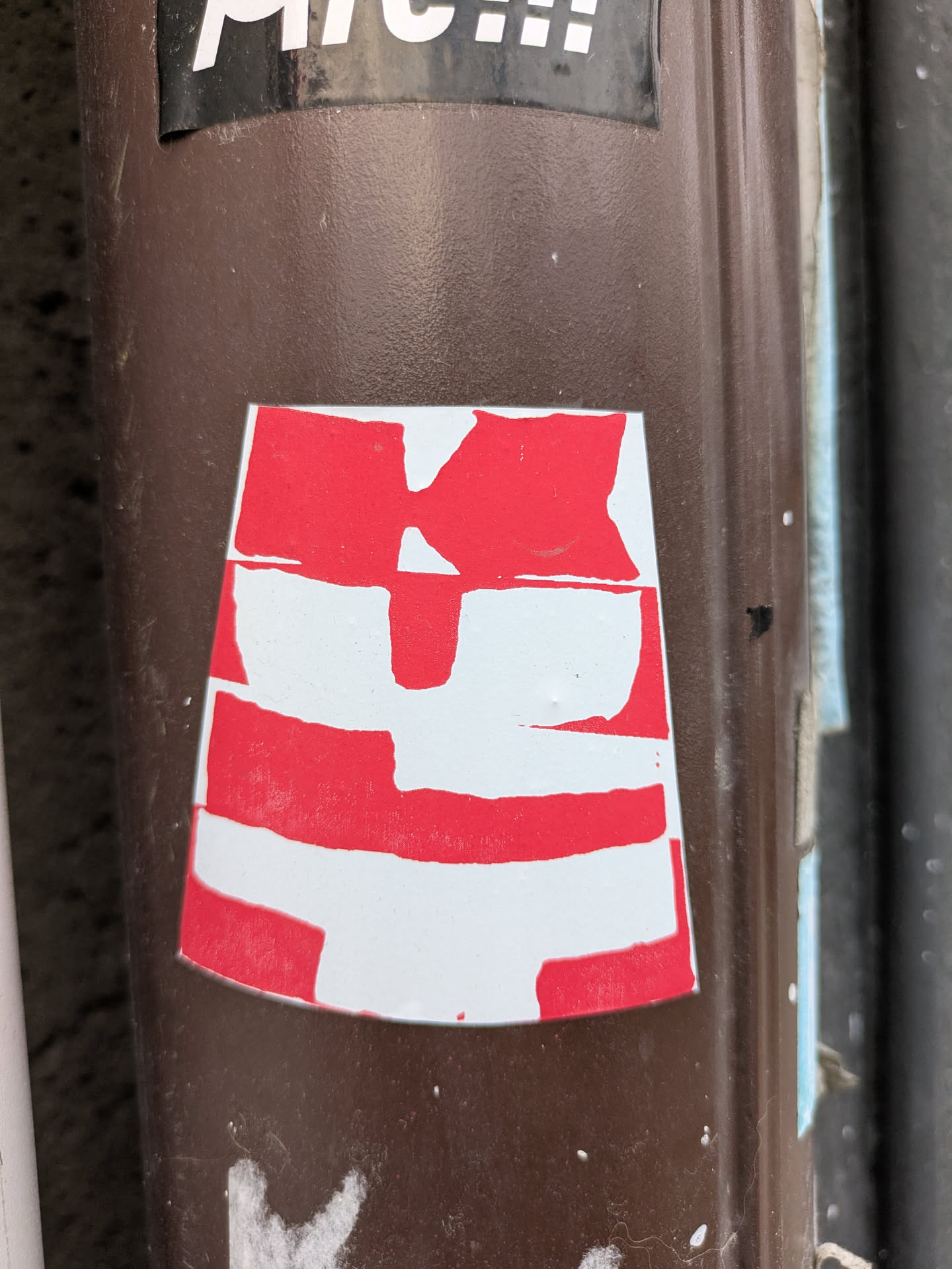 A close-up shot of a red and white abstract sticker on a brown surface. The sticker appears to be hand-made or screen-printed.  There is no discernible text or QR code.