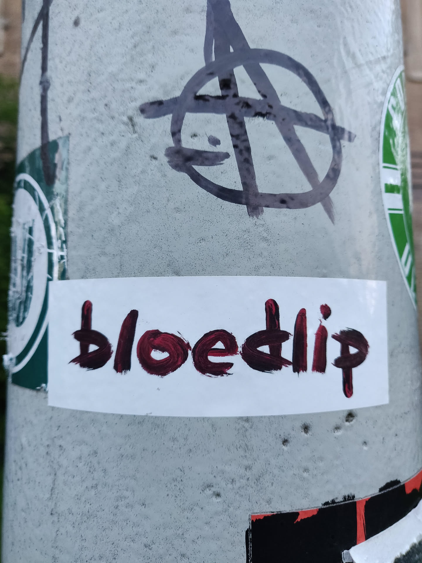 A white rectangular sticker with the word \"bloedip\" written in dark red paint. The lettering style is slightly irregular, giving it a hand-painted look.  The sticker is affixed to a gray pole, which also has other graffiti and stickers on it.