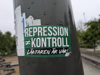A rectangular sticker adhered to a dark grey pole, showing signs of wear and water droplets. The main body of the sticker has a vibrant green background. The top two lines of text are in large, bold white capital letters: 'REPRESSION' followed by a 'not equal to' symbol and 'KONTROLL'. Below this, there's a black horizontal bar with smaller, white, italicized text that reads 'LÄKTAREN ÄR VÅR!'. The sticker appears slightly crumpled and worn, particularly at the edges, and there's a partially visible, torn white and blue sticker underneath it on the left side.