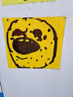 This sticker is square and yellow, with a dark brown design reminiscent of a face or possibly a stylized creature.  The brown paint or ink appears to have some texture or glitter to it. The style is playful and slightly abstract.