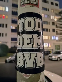 A close-up shot of a dark blue sticker with white block lettering that reads "TODDEM BYB". The sticker is affixed to a lamppost in an urban environment at night.  The background is slightly blurry, showcasing buildings and parked cars. 