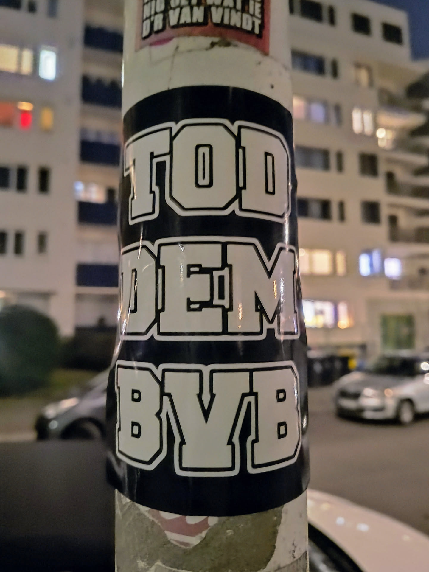 A close-up shot of a dark blue sticker with white block lettering that reads "TODDEM BYB". The sticker is affixed to a lamppost in an urban environment at night.  The background is slightly blurry, showcasing buildings and parked cars. 