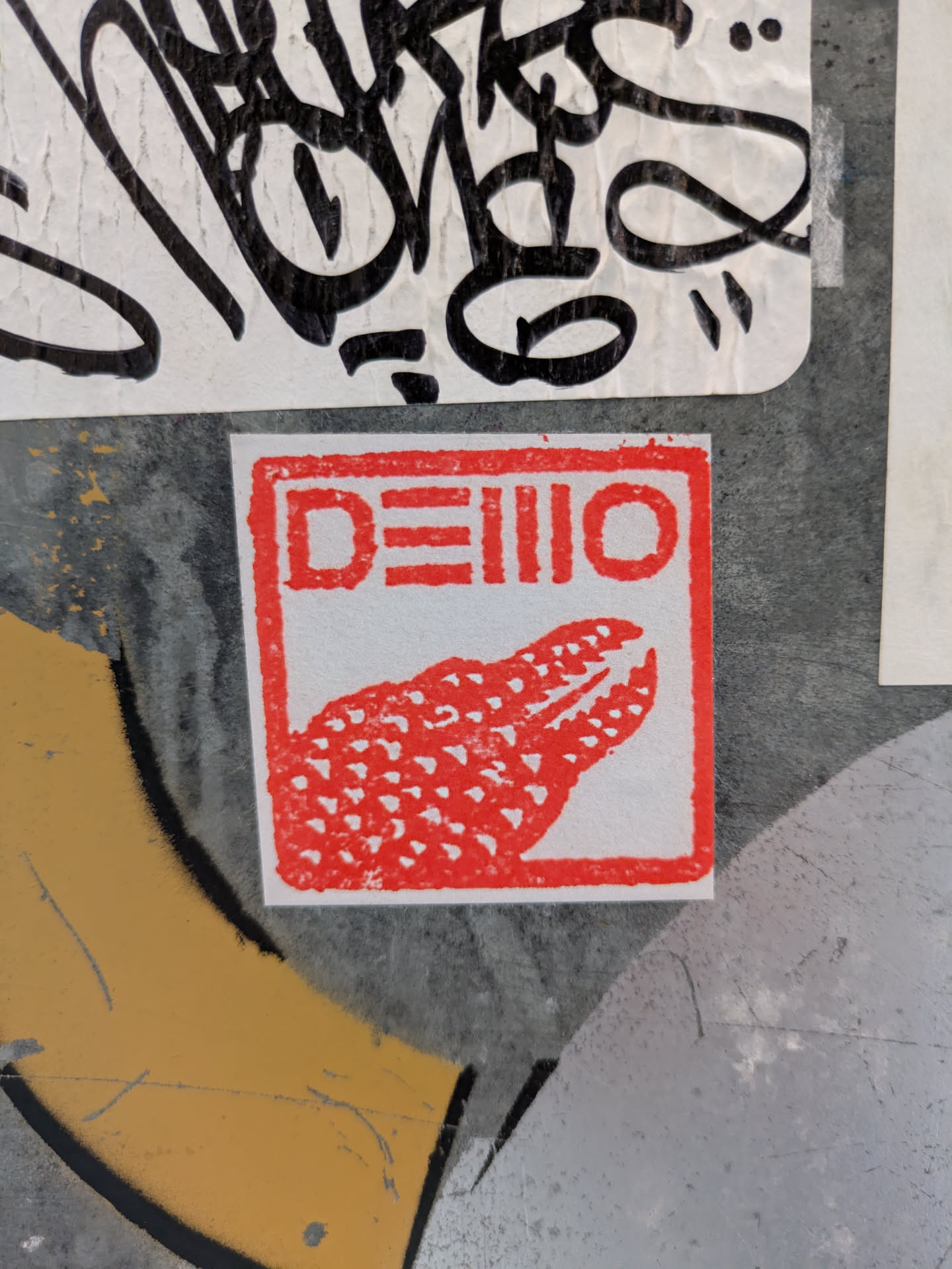 A red square sticker with a stylized alligator and the text DEIII0 is affixed to a wall. The sticker appears to be hand-printed or stamped, giving it a slightly rough texture.
