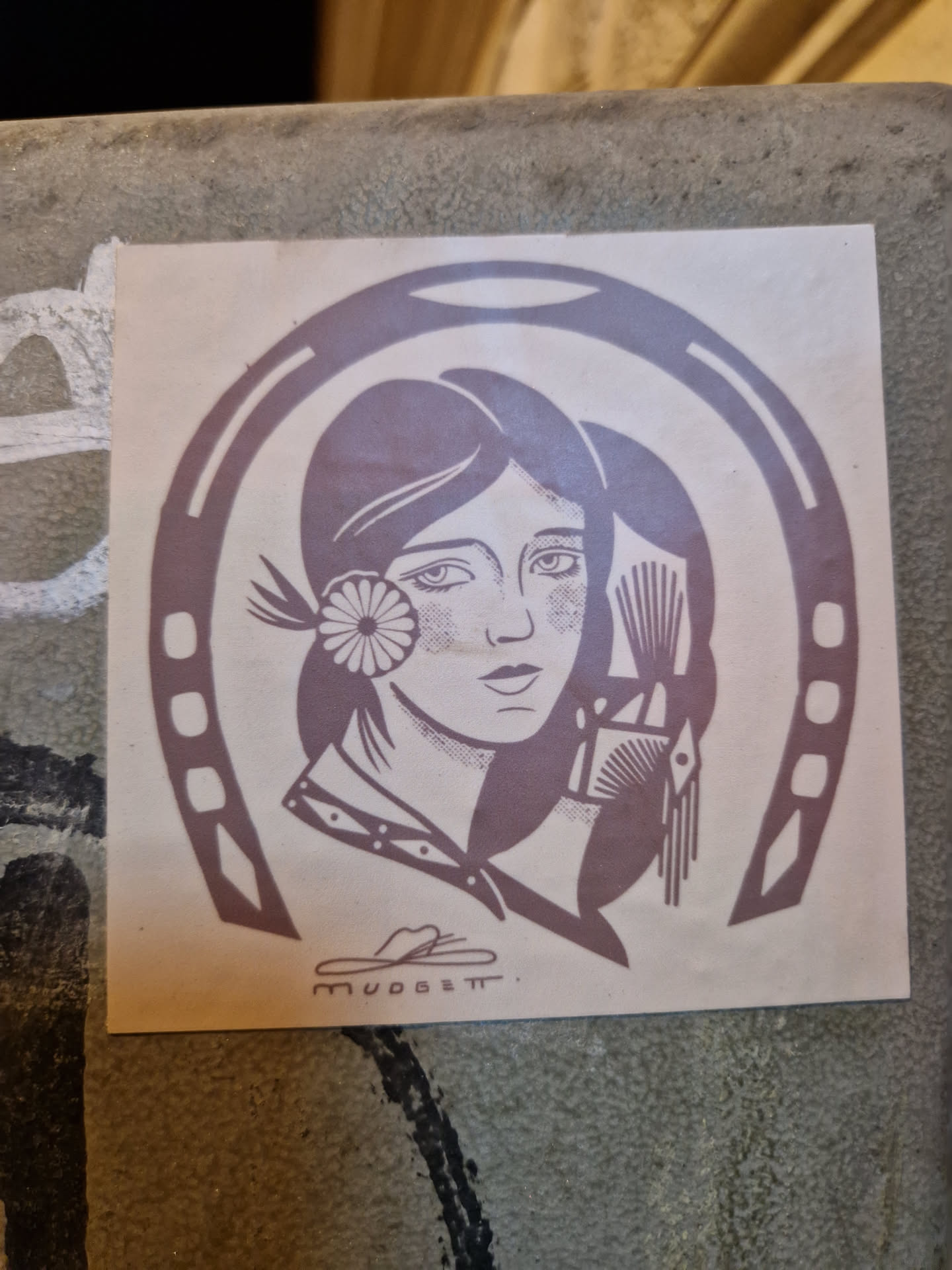 A sticker featuring a vintage illustration of a woman's face inside a horseshoe. The style is reminiscent of Art Deco or Art Nouveau. The sticker is in muted purplish-brown tones, and the artist's signature is visible in the bottom-center of the design. 