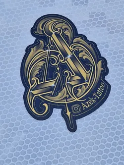 A stylish sticker featuring the Azek Tattoo logo. The logo is a gold and black design with intricate details, giving it a luxurious feel.  The 'A' is prominently featured, incorporated within decorative flourishes.  The sticker is likely used for branding and promotion by the tattoo studio.