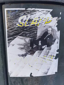 A sticker featuring a black and white photo of a man sitting on some stairs, with the words "SLAP" and "KOMUNIST" overlaid in yellow and blue. The sticker is partially torn and weathered.