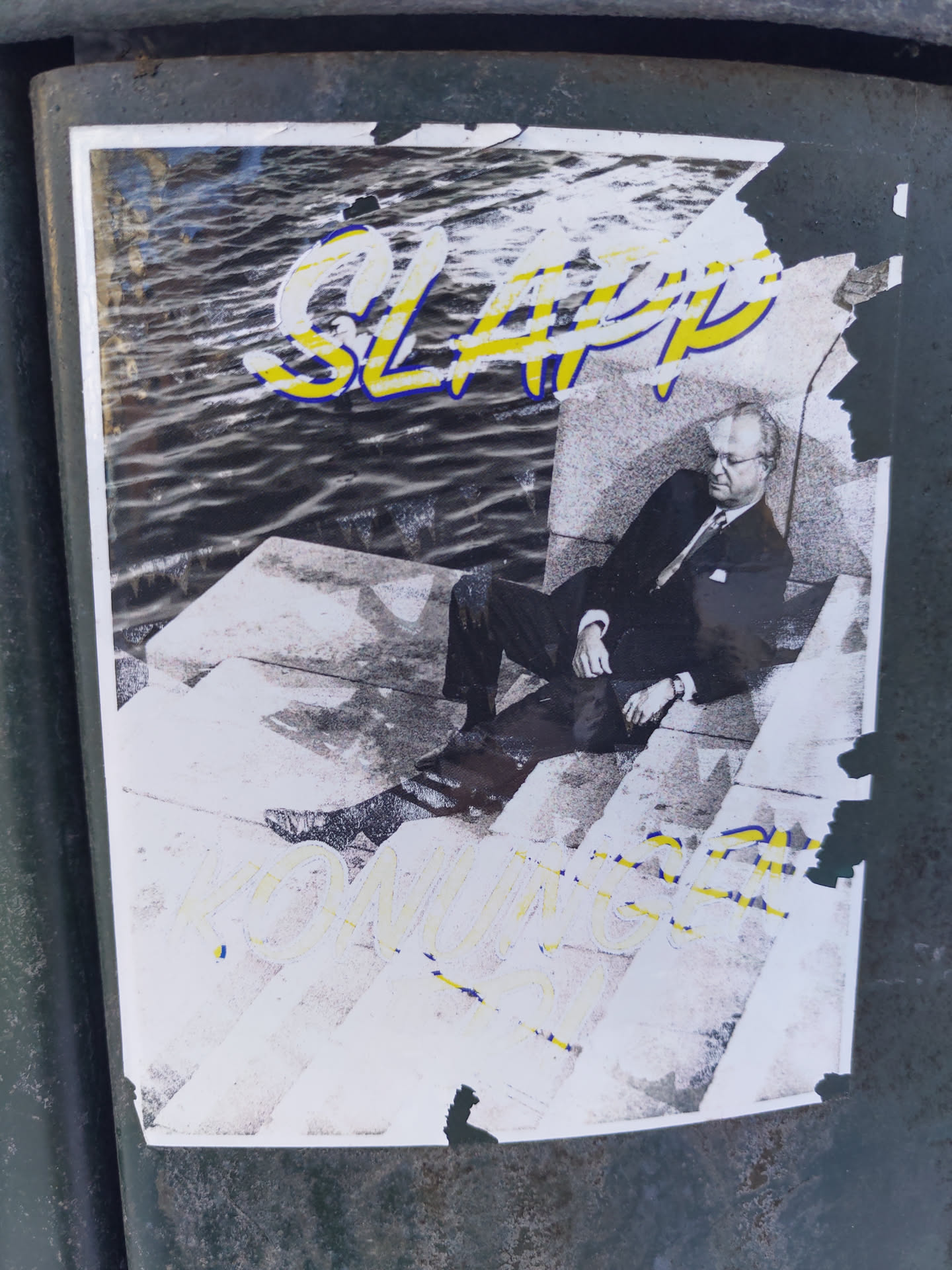 A sticker featuring a black and white photo of a man sitting on some stairs, with the words "SLAP" and "KOMUNIST" overlaid in yellow and blue. The sticker is partially torn and weathered.