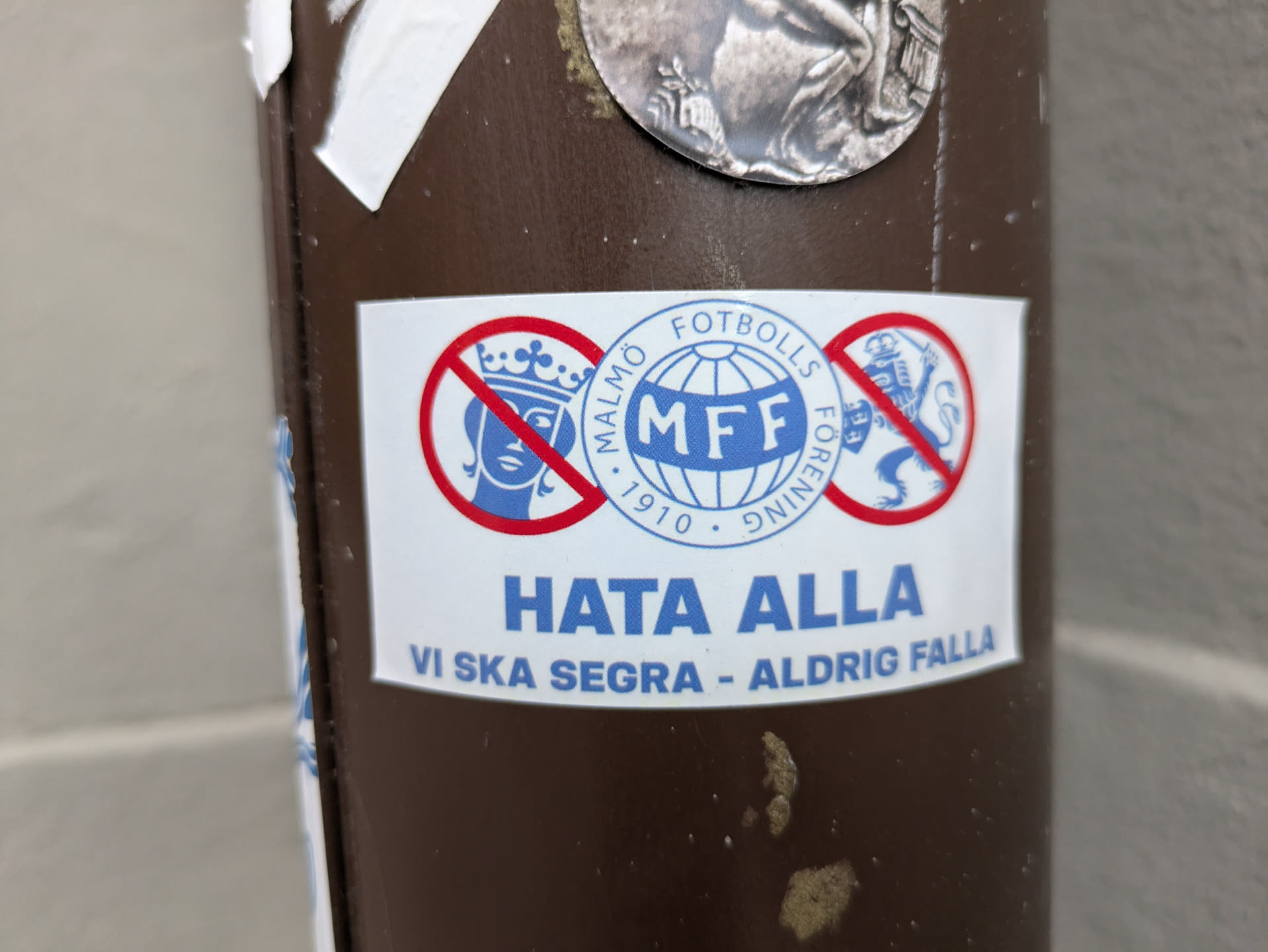 A sticker expressing loyalty to Malmö Fotbollsförening (MFF).  It features a prohibition symbol over the coat of arms of Sweden and another emblem, with the text "Hata Alla" (Hate All) and "Vi ska segara - Aldrig falla" (We shall conquer - Never fall).