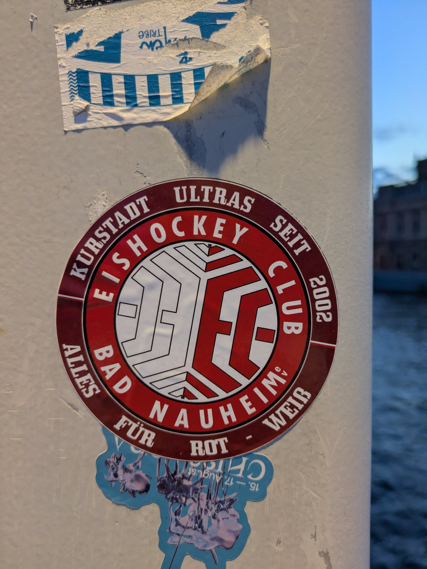 The sticker is circular with a dark red border. Inside the border, text reads 'KURSTADT ULTRAS EISHOCKEY CLUB SEIT 2002 ALLES FÜR BAD NAUHEIM ROT - WEISS'. The center features a stylized logo with the letters 'ECC' formed from white lines against a red background, with a geometric, paneled design. Below this main sticker, part of another sticker is visible, featuring a light blue background with metallic-looking figures and text that appears to be a date.