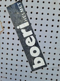 A weathered sticker advertising a Poeri Helmet. The sticker is black with white lettering and is affixed to a perforated grey surface. The text 'Poeri' is prominently displayed, with 'Helmet' written vertically alongside it.