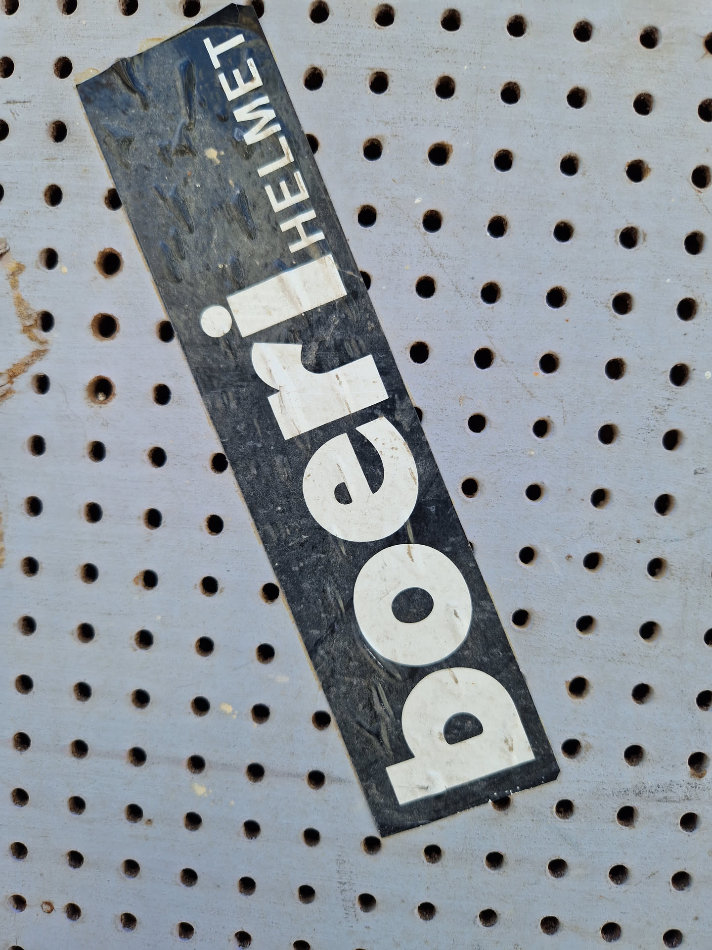 A weathered sticker advertising a Poeri Helmet. The sticker is black with white lettering and is affixed to a perforated grey surface. The text 'Poeri' is prominently displayed, with 'Helmet' written vertically alongside it.