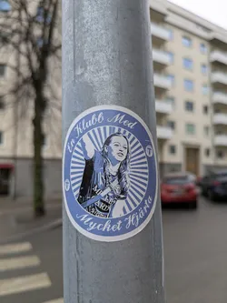 This is a round, blue and white sticker that is adhered to a pole. The design features a woman with text around her that reads "En Klubb Med Mycket Hjarta". The woman is wearing a sports shirt and jacket.