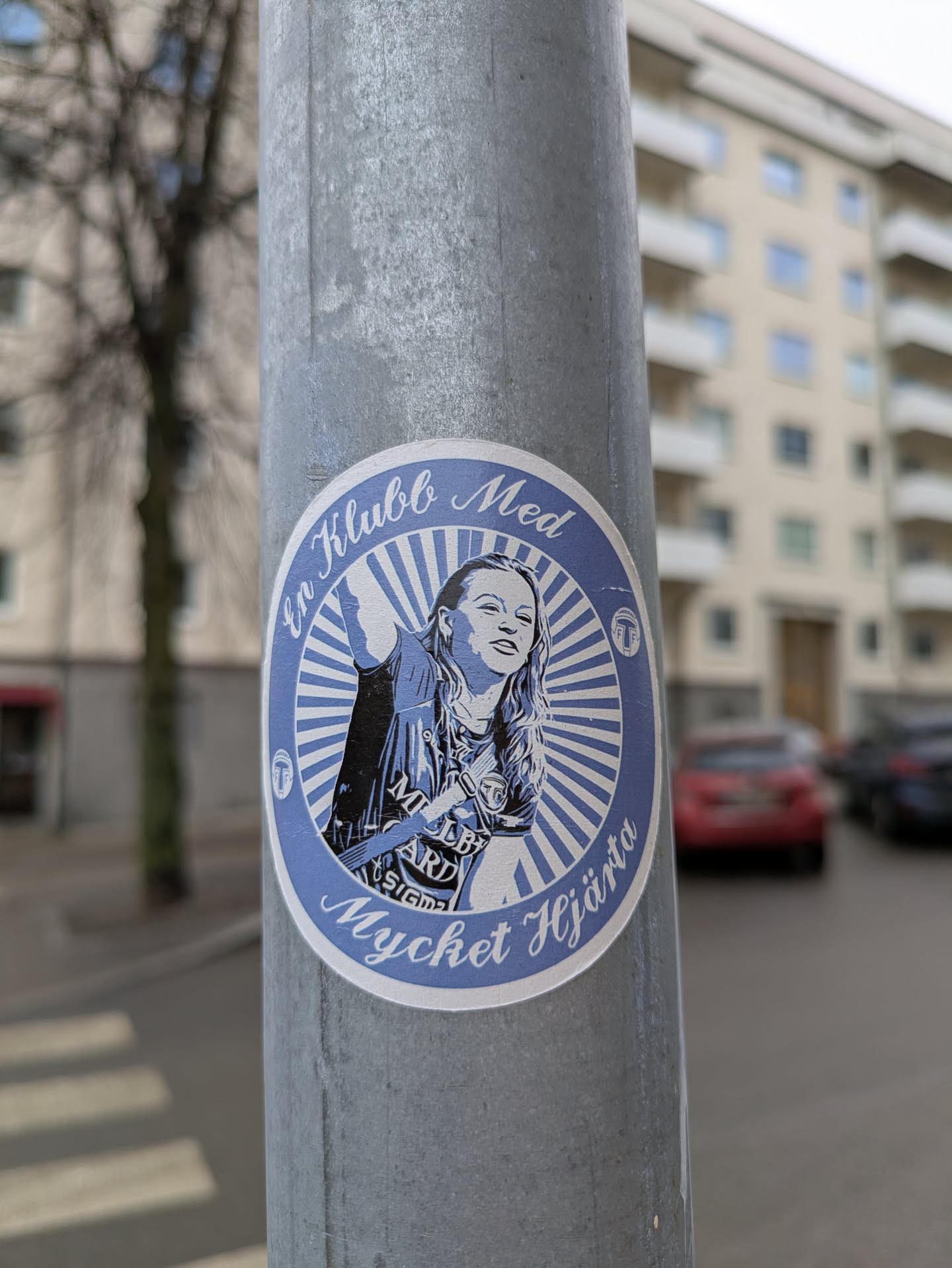 This is a round, blue and white sticker that is adhered to a pole. The design features a woman with text around her that reads "En Klubb Med Mycket Hjarta". The woman is wearing a sports shirt and jacket.