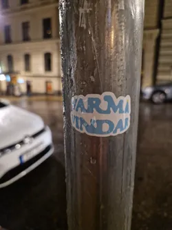 A light blue sticker with the words "VARMA VINDAR" in a stylized font.  The sticker is affixed to a metal lamppost outside at night. The background is blurred, showing a city street and cars.