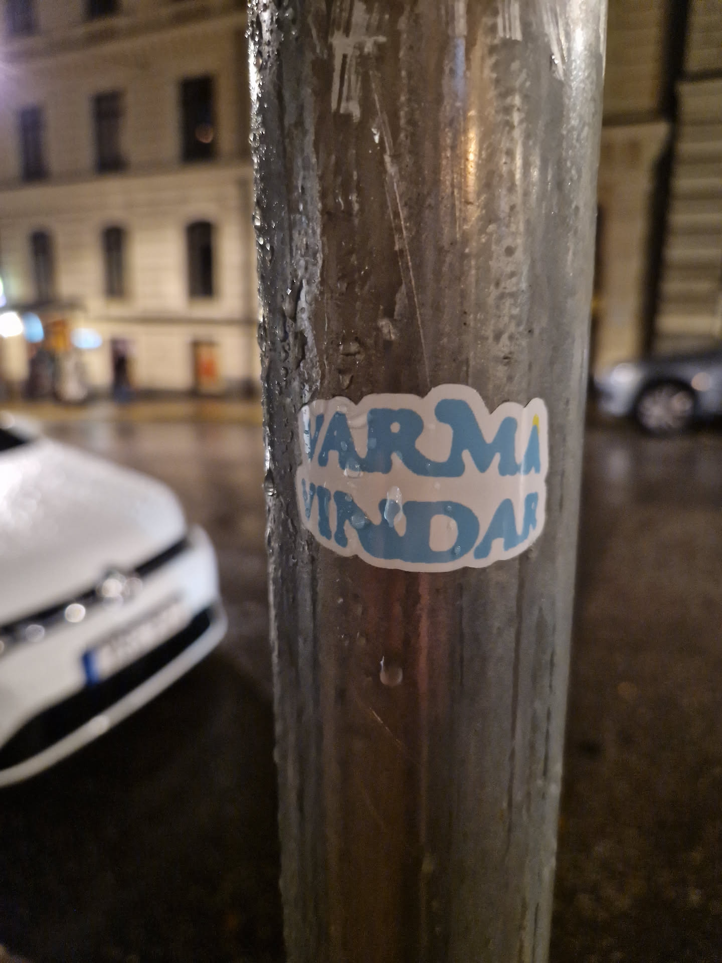A light blue sticker with the words "VARMA VINDAR" in a stylized font.  The sticker is affixed to a metal lamppost outside at night. The background is blurred, showing a city street and cars.