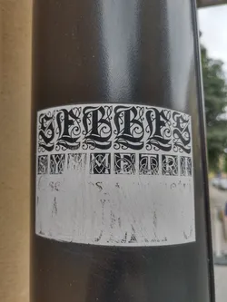 Black and white sticker with a symmetrical design. The word SLEBBLES is written in a stylized font at the top, and a line of smaller, symmetrical letters is below it. There is a blank space at the bottom that seems to be intended for text or other information. The sticker is affixed to a dark gray pole.