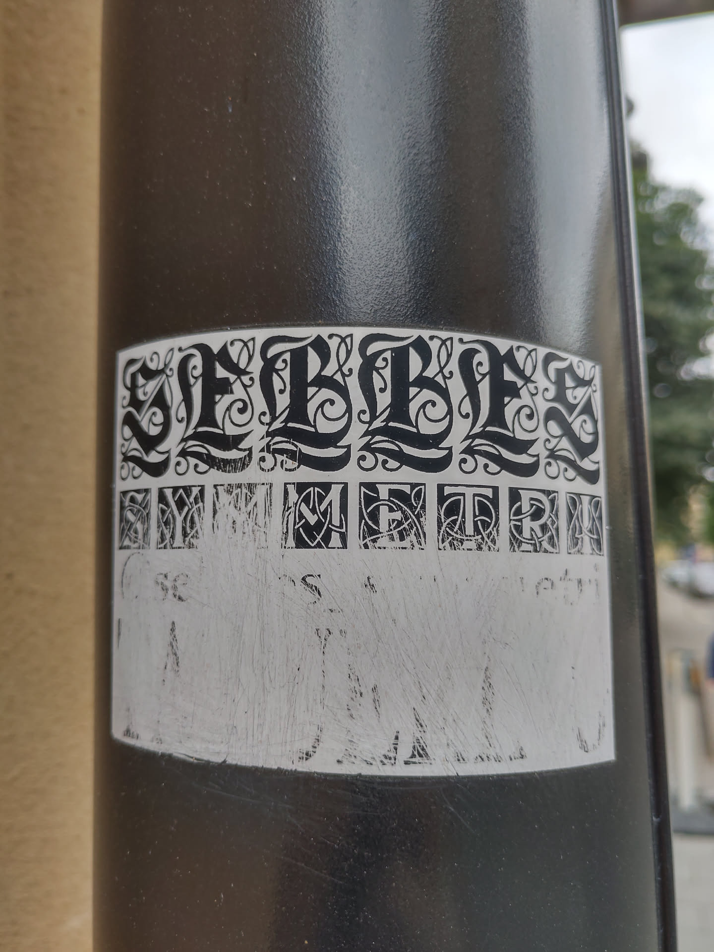 Black and white sticker with a symmetrical design. The word SLEBBLES is written in a stylized font at the top, and a line of smaller, symmetrical letters is below it. There is a blank space at the bottom that seems to be intended for text or other information. The sticker is affixed to a dark gray pole.
