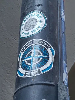 A Stone Island On Tour sticker on a pole. The sticker is blue and white with the Stone Island logo. The text "ON TOUR" is written below the logo. There is another sticker on top of it with text "CRUISERS" and a ball image.