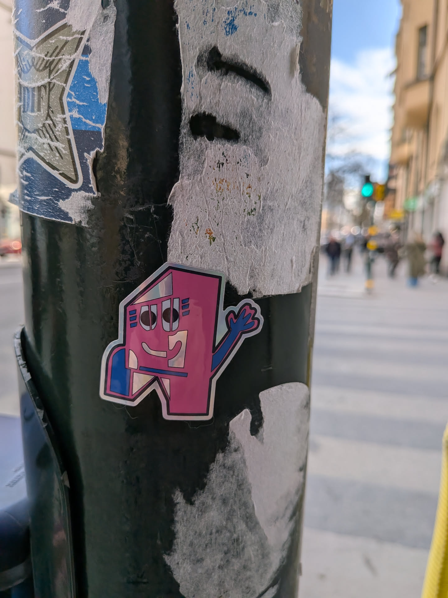 The sticker depicts a vibrant, cartoonish character with a geometric, house-like or blocky 'A' shape as its body. It is predominantly magenta/pink with bold black outlines. The character has a friendly face featuring two large, round black eyes with white pupils and a wide, curved smile. Three small blue rectangular accents are placed on either side of its head. A blue arm extends from its right side, bent at the elbow, with a four-fingered hand waving. A blue block-like element is visible on its lower left. The sticker has a glossy finish, with reflective, possibly holographic elements visible within the pink and blue areas, giving it a metallic sheen. It is cleanly die-cut and applied to a dark green pole.