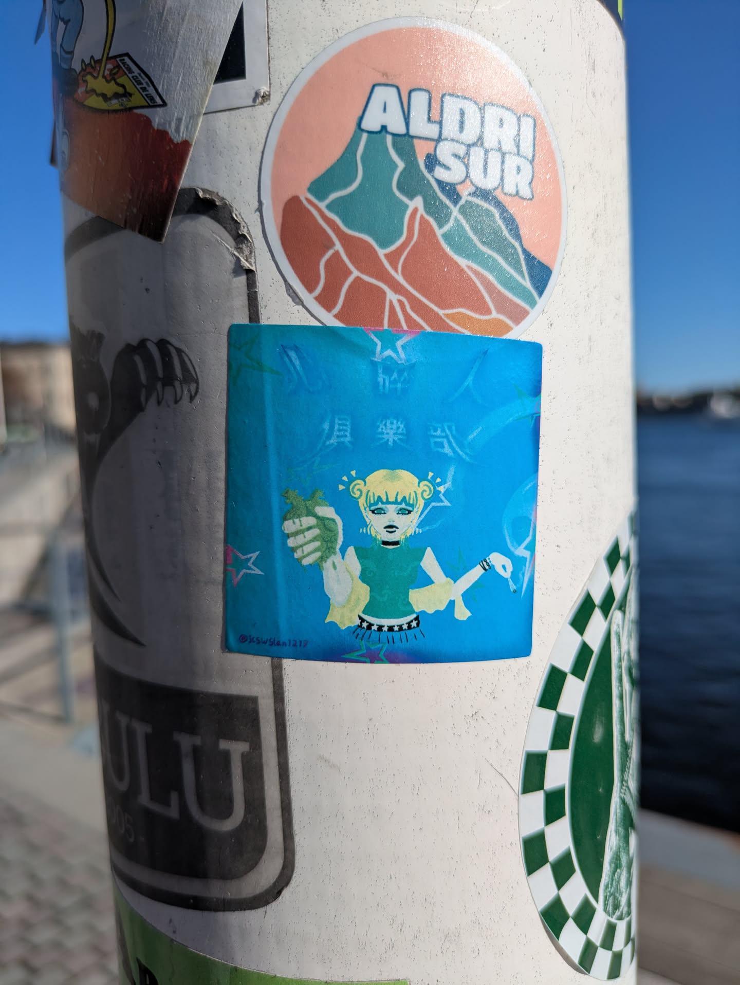 A blue square sticker on a light pole features a girl with blonde hair, dressed in green, holding money in one hand and a cigarette in the other. The text above the girl reads '俱樂部式'. The background is a light blue with stars.