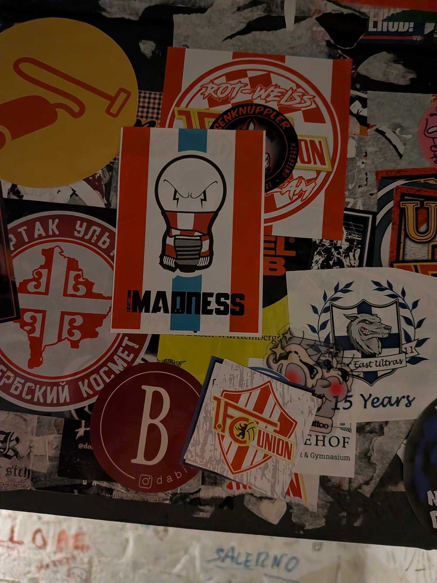 A vertically oriented rectangular sticker featuring a cartoon lightbulb character with a fierce, scowling facial expression. The lightbulb wears a red and white striped supporters' scarf wrapped around its neck just above the metal screw base. The background features vertical stripes of orange-red, white, and a central light-blue stripe. At the bottom, the word 'MADNESS' is displayed in bold black lettering, preceded on the left by the vertically printed word 'LIGHTBULB'.