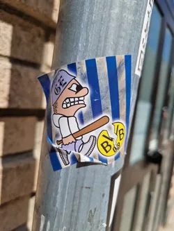 The sticker features a cartoon illustration of an angry-looking character wearing a cap, swinging a baseball bat. The background is blue and white striped. A small circular logo is partially visible next to the character. The art style is reminiscent of comic books.