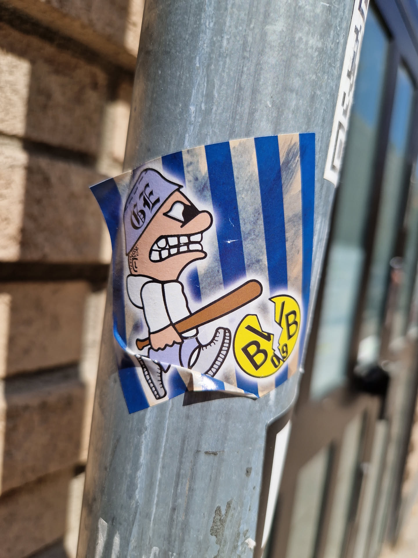 The sticker features a cartoon illustration of an angry-looking character wearing a cap, swinging a baseball bat. The background is blue and white striped. A small circular logo is partially visible next to the character. The art style is reminiscent of comic books.