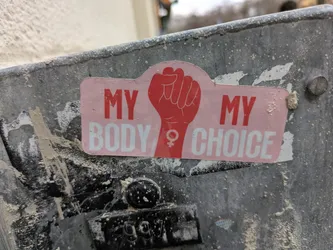 Pink and red sticker with a raised fist and the text "My Body My Choice". The sticker is affixed to a dirty gray metal surface.