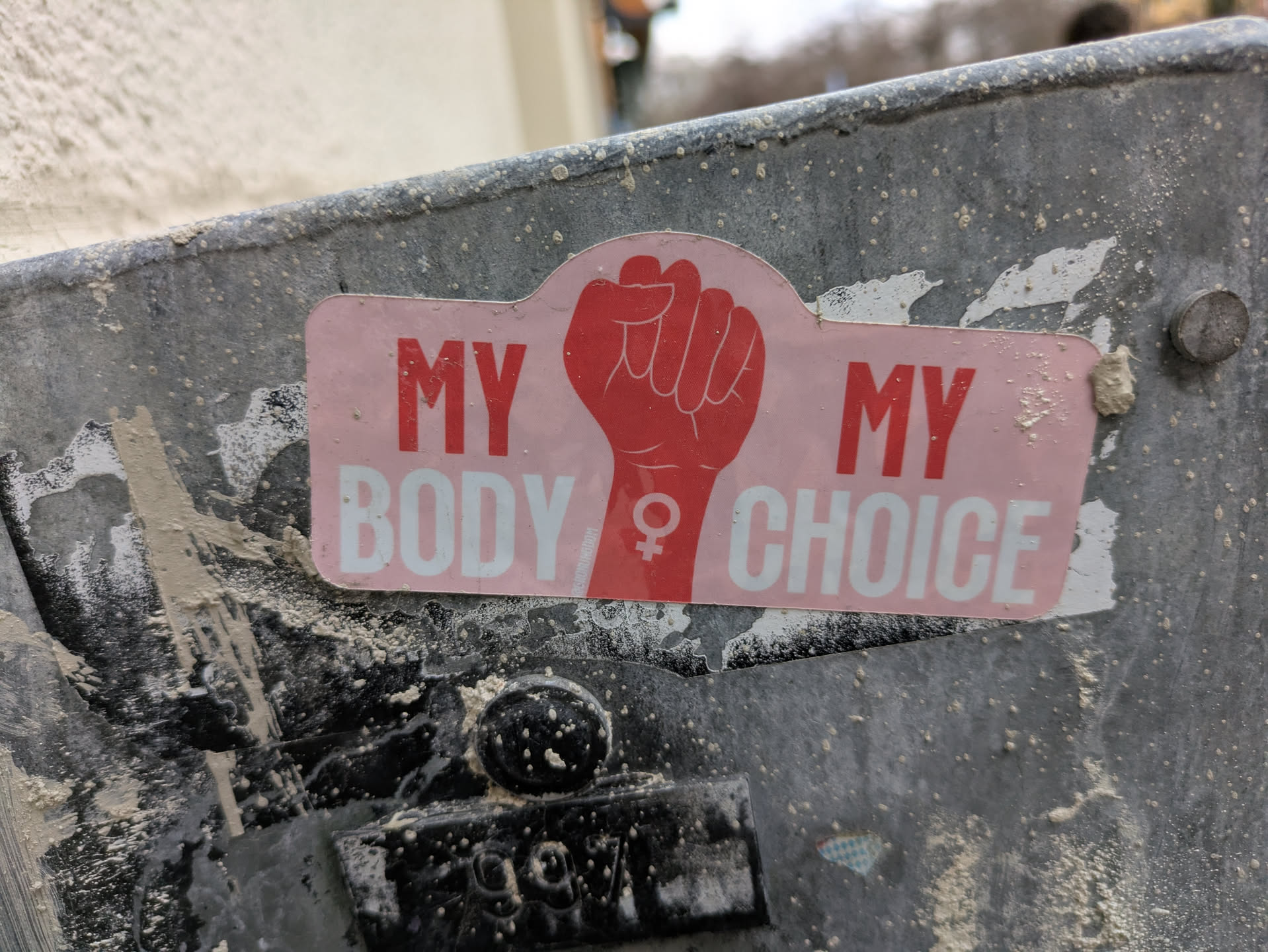 Pink and red sticker with a raised fist and the text "My Body My Choice". The sticker is affixed to a dirty gray metal surface.