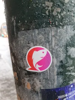 A vibrant pink and purple gradient sticker featuring a stylized fish design. The fish is depicted in a circular shape, creating a visually appealing emblem.  The sticker appears to be affixed to a weathered pole outdoors.
