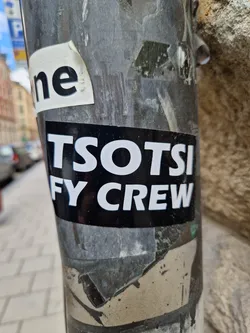 A black and white sticker with the text "TSOTSI FY CREW" on a dark background. It is affixed to a pole, which also has other stickers and debris.