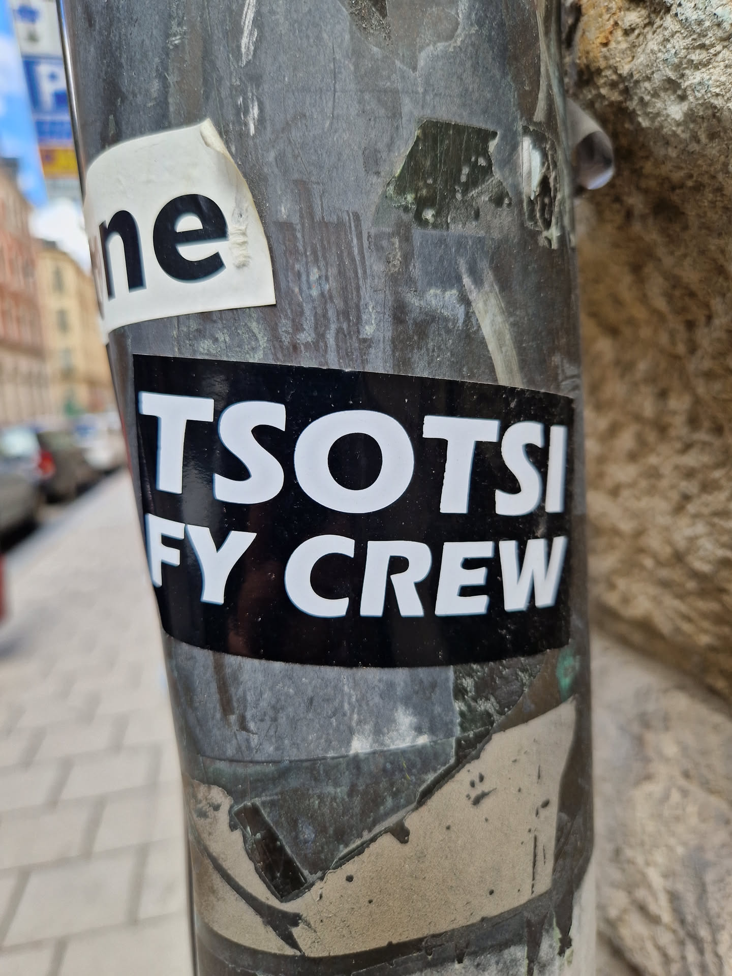 A black and white sticker with the text "TSOTSI FY CREW" on a dark background. It is affixed to a pole, which also has other stickers and debris.