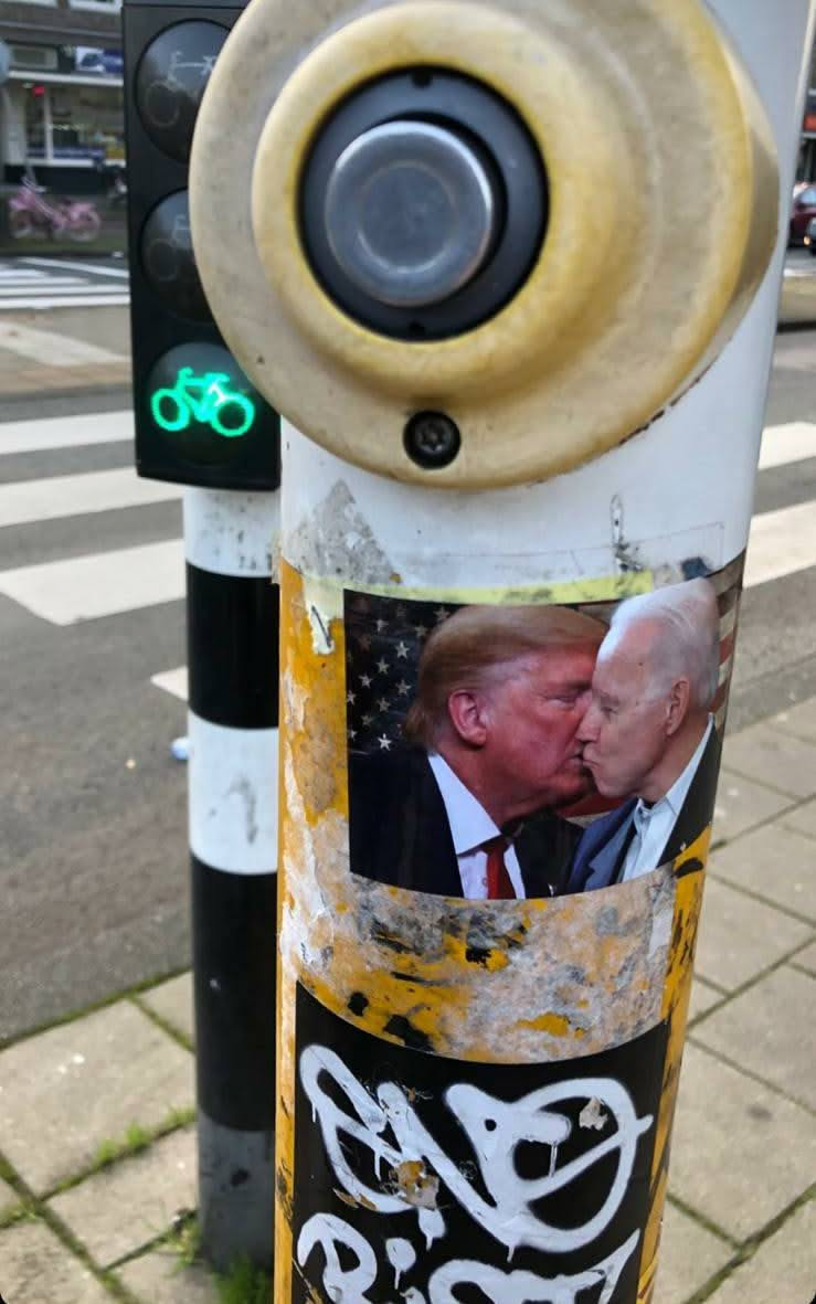 A sticker depicting Donald Trump and Joe Biden kissing.  The sticker is affixed to a traffic pole, showing some wear and tear.