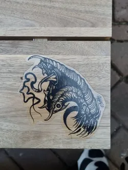 Close-up of a black and white sticker featuring a detailed illustration of a claw, possibly belonging to a mythical creature or a dragon. The style is intricate and detailed, with sharp lines and shading to create depth and texture. The sticker is affixed to a light brown wooden surface.