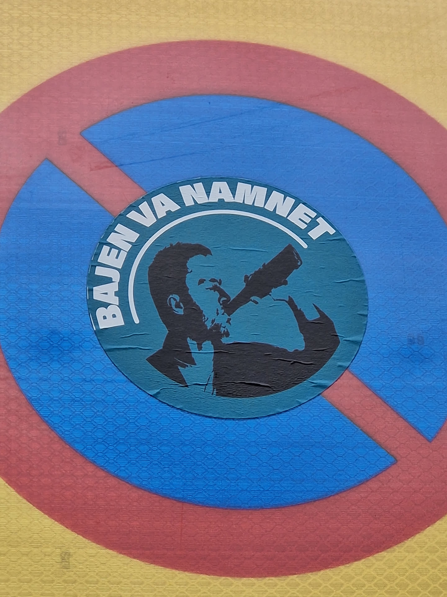 A sticker featuring a silhouette of a man drinking from a bottle, set within concentric circles of red, blue and yellow. The text 'Bajen va namnet' is visible around the central image.